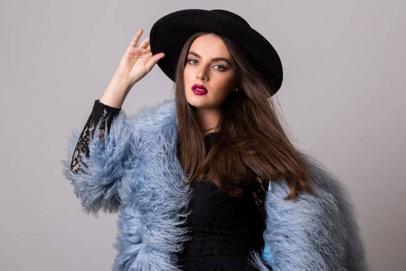 Studio fashion portrait of gorgeous  woman in stylish winter fluffy  blue coat and black hat posing on bright  grey background.